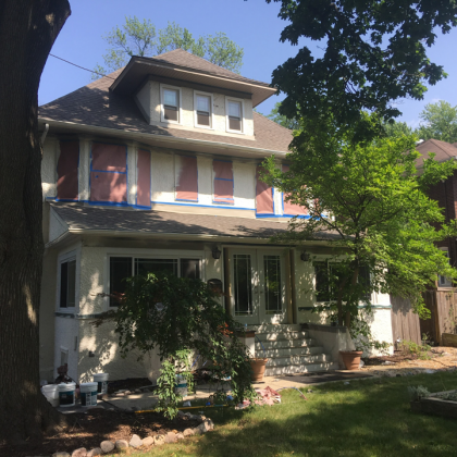 Two-story house undergoing renovation; windows covered, stucco exterior, front porch, green lawn, trees, blue sky.