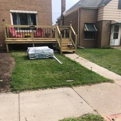 Newly built wooden deck with stairs and stacked lumber on the grass next to a sidewalk and brick house.