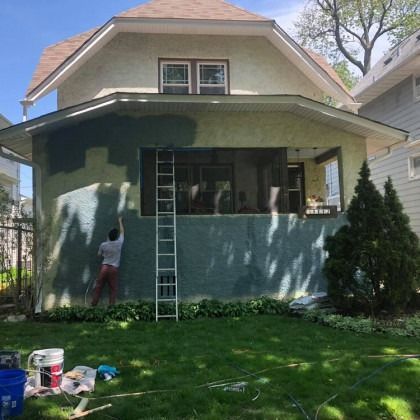 Person painting a house exterior, working from a ladder. Green house with a green porch, blue sky.