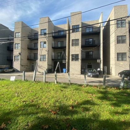 Apartment building with balconies, beige brick facade, and green grass in front. Cars parked on the side.