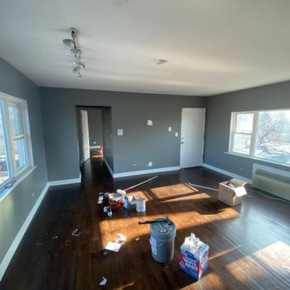 Empty room with dark wood floors and gray walls. Paint supplies and a door are visible. Sunlight.