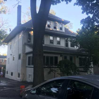 Three-story house with white stucco walls, windows, and a tall tree in front; a car is parked nearby.