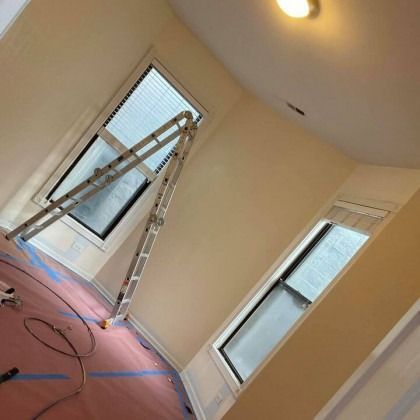 A ladder leaning against a window in a room being painted, with two windows and protective floor covering.
