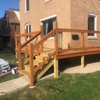 Wooden deck with steps attached to a brick house. Railings are stained brown, with green grass in the foreground.