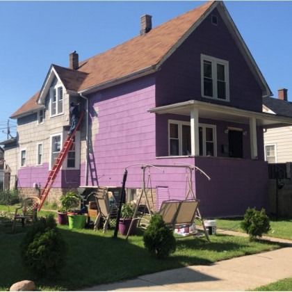 House being painted; one side purple, one side gray. Person on ladder, brown roof, yard with lawn furniture.