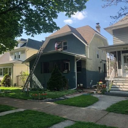 Person painting the exterior of a two-story house, using a ladder. The house has blue siding and a brown roof.