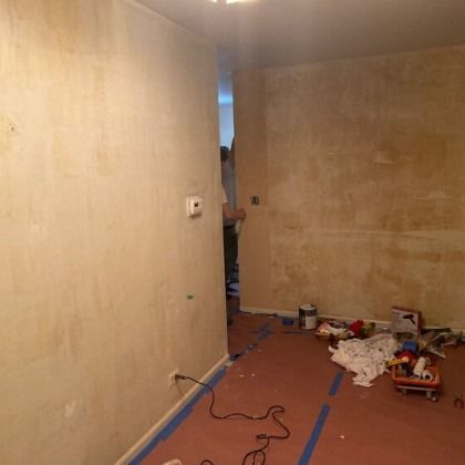 Room with exposed walls, red floor, blue tape, and construction debris. Someone is visible in doorway.