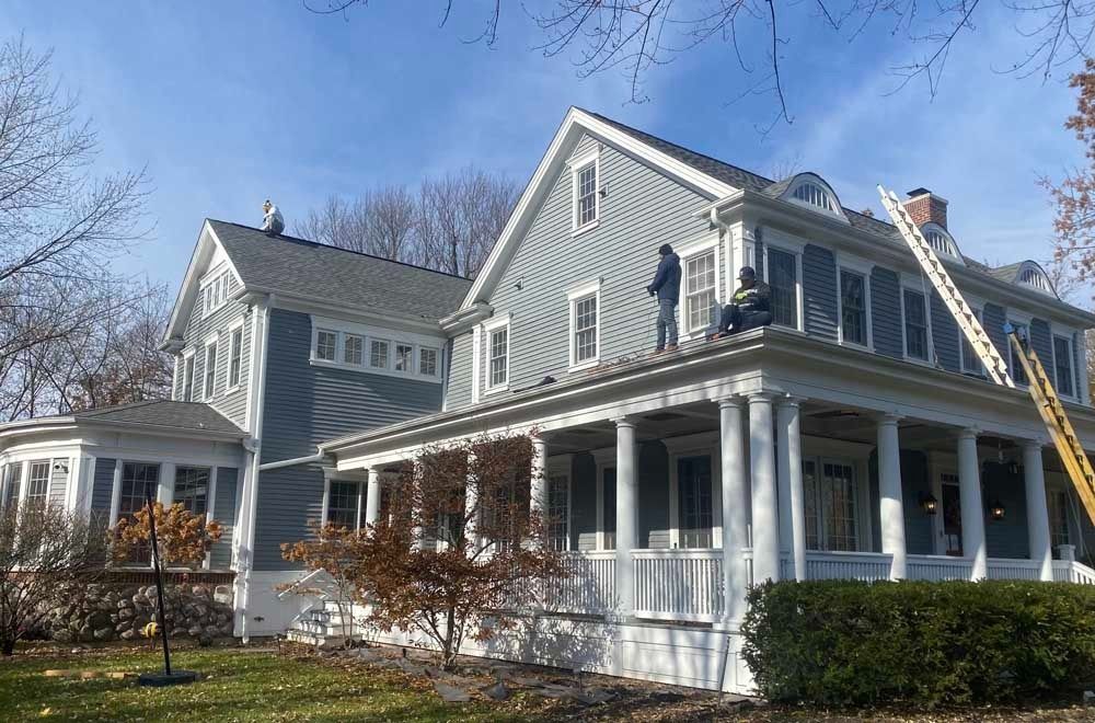 Workers on a two-story house with light blue siding, white trim, and a porch, under a blue sky.