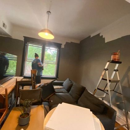 Person painting a room with a dark green wall, gray couch, and a ladder.