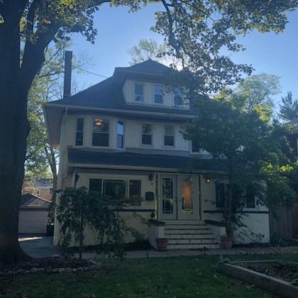 Two-story house with light-colored siding, dark roof, chimney, front steps, and a lush green yard with trees.