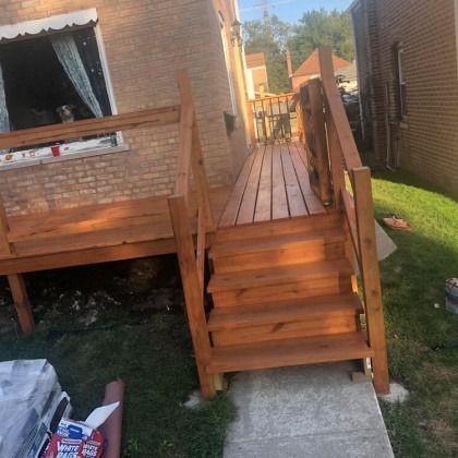 Wooden deck with stairs, stained brown, attached to a brick building, set on a green lawn.