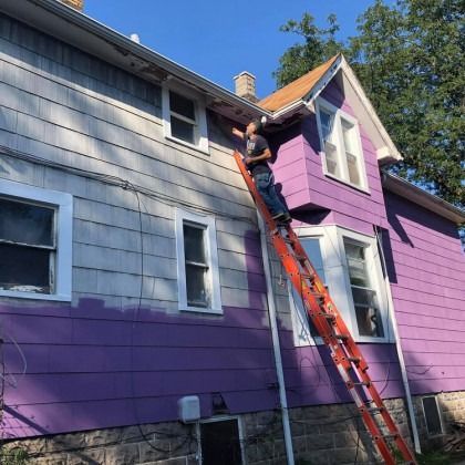 Person on a ladder painting a purple house; sky and trees visible.