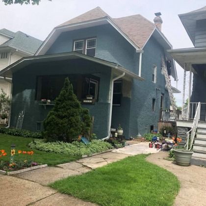 Two-story teal house with brown roof and ladder propped against the side, porch and small garden in front.