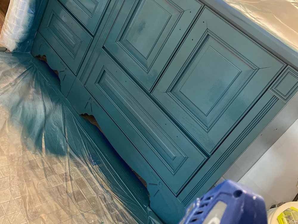 Teal painted wooden cabinet being sprayed with a paint sprayer, covered with plastic sheeting.