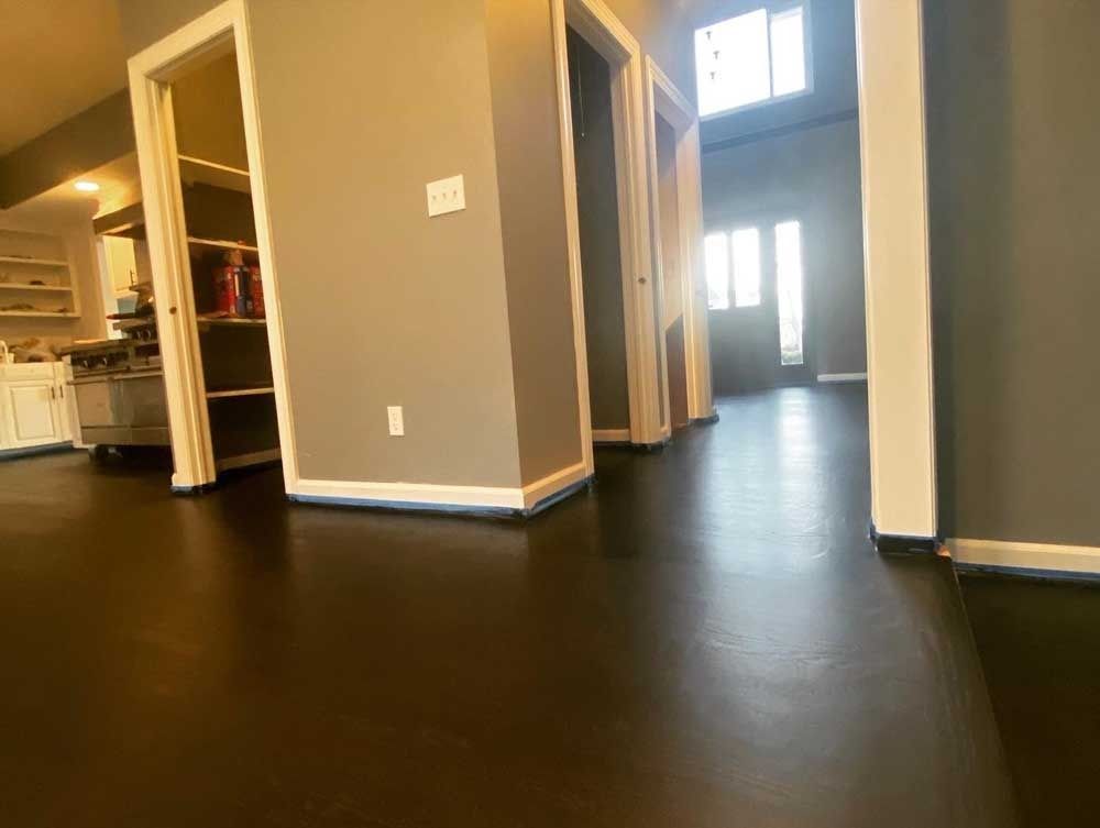 Dark brown floor in a hallway with gray walls and doorways, leading to a front door with light.