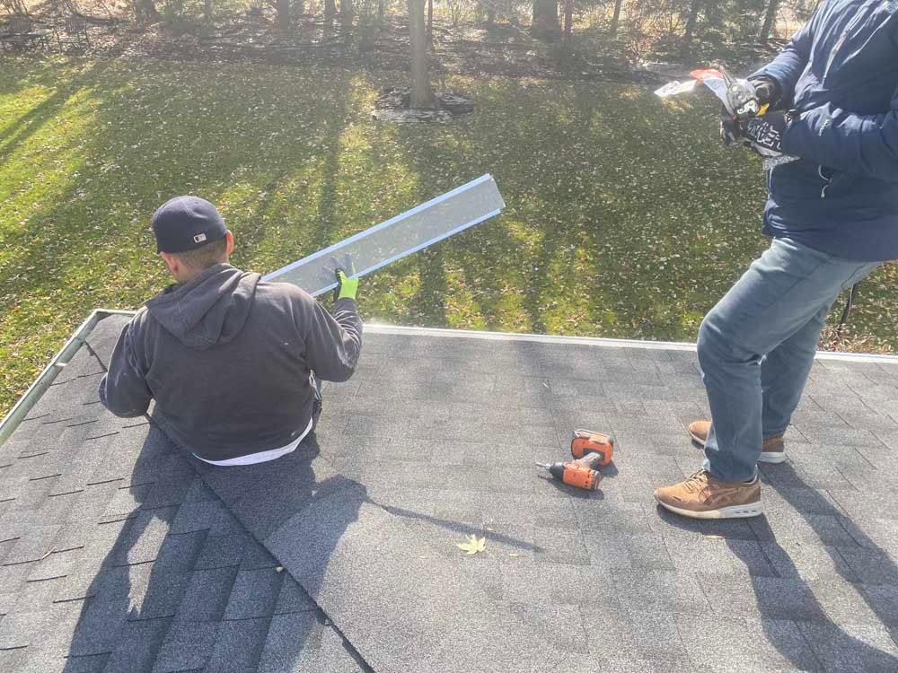 Two workers on a roof, installing a silver flashing. One kneels holding the material, the other stands.