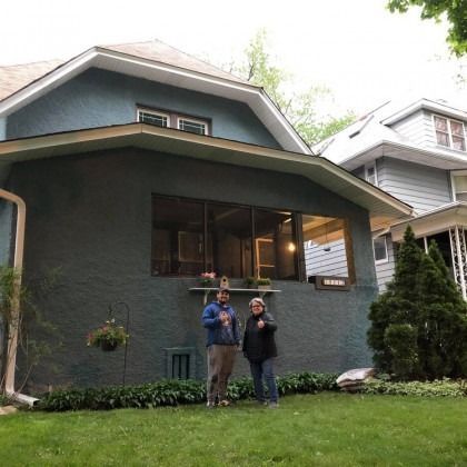 Two people stand in front of a blue-painted house with large windows. One gives a thumbs-up.