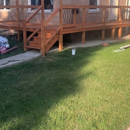 Wooden deck with stairs, stained brown, on a grassy lawn; a brick building is behind it.