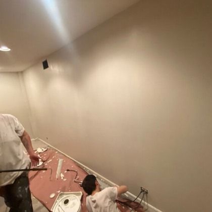 Two people painting an interior wall. One is standing, the other is kneeling near electrical wires. The wall is light.