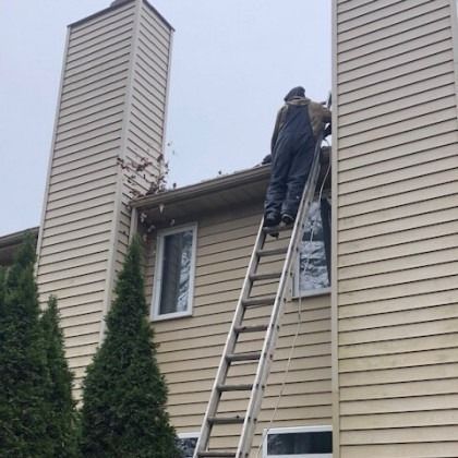 Person on a ladder cleaning gutters on a two-story house with two chimneys and siding; cloudy sky.