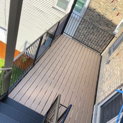 Elevated deck with tan composite boards, black railing, and black metal stairs. Attached to brick building.