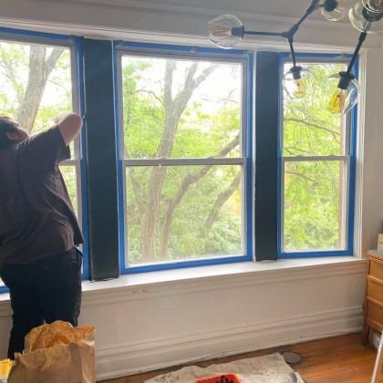 Person taping window frames before painting, trees visible outside.