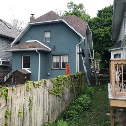 Blue house with brown roof, white trim. Ladder against side. Wooden fence with ivy.