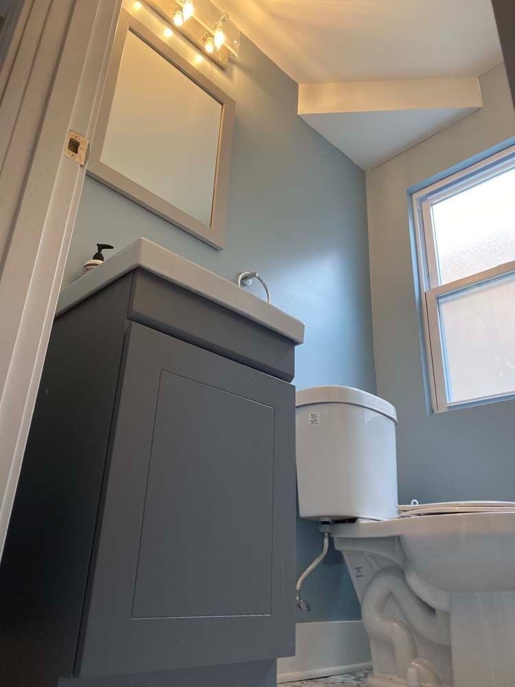 A small bathroom with a blue wall, gray vanity, toilet, mirror, and a window.