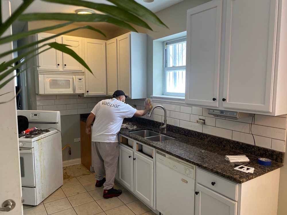 Man cleaning a kitchen counter with white cabinets and a window above a sink.