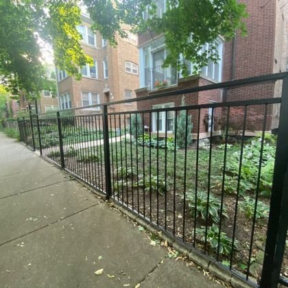 Black metal fence in front of a small garden with plants, bordering a sidewalk, with a brick building in the background.