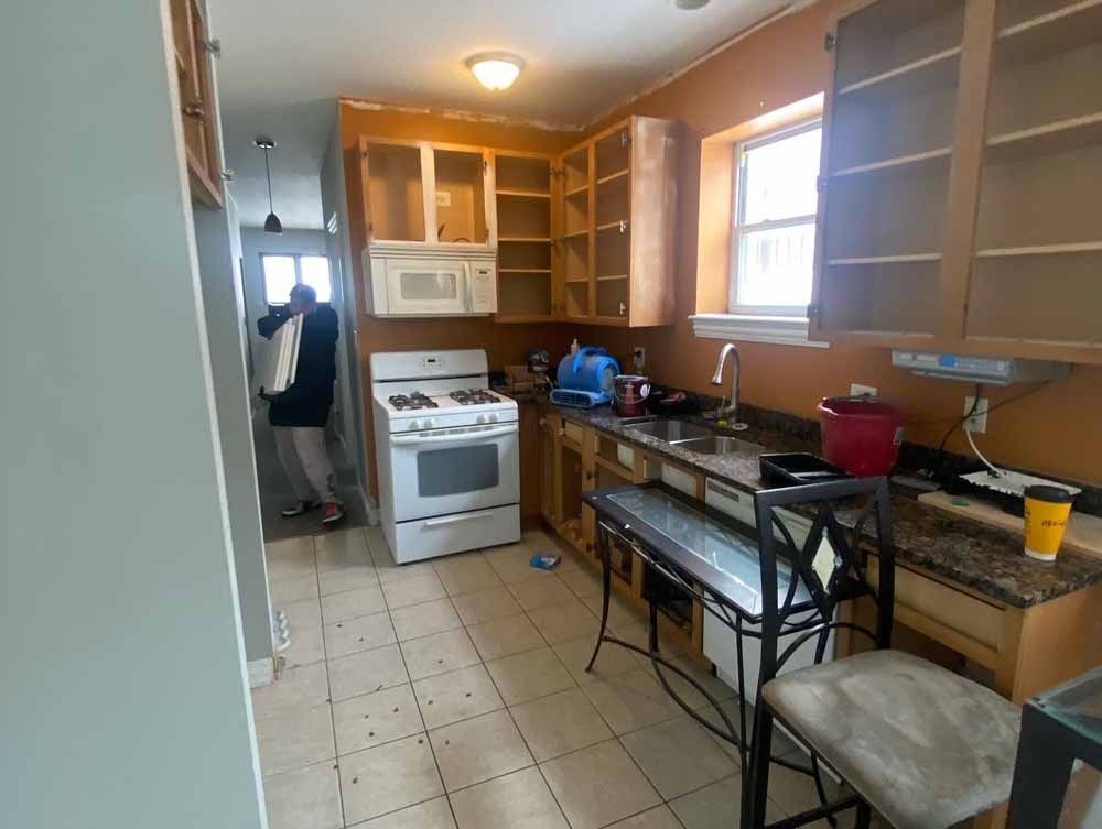 Kitchen renovation in progress: bare cabinets, appliances, small window, person in doorway.