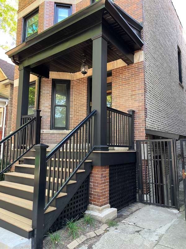 Brick building with a covered porch, stairs, and black railing. Exterior view, gray concrete below.