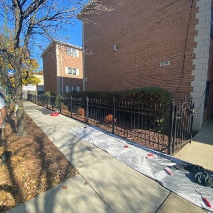Black metal fence borders a sidewalk next to a brick building; leaves and sidewalk debris are present.