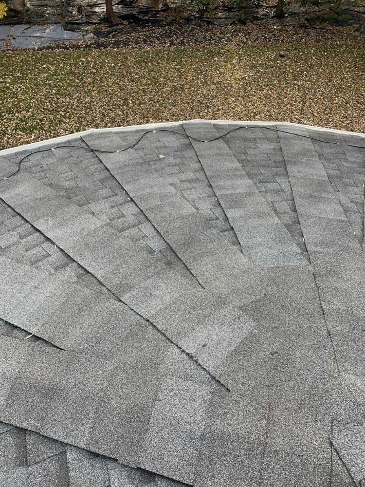 Close-up of a weathered asphalt shingle roof. Gray shingles show cracks and wear. Background is a yard with brown grass.