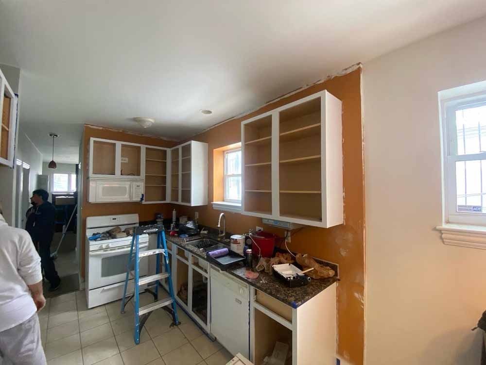 Kitchen undergoing renovation: white cabinets, orange walls, appliances, ladder, and two people.