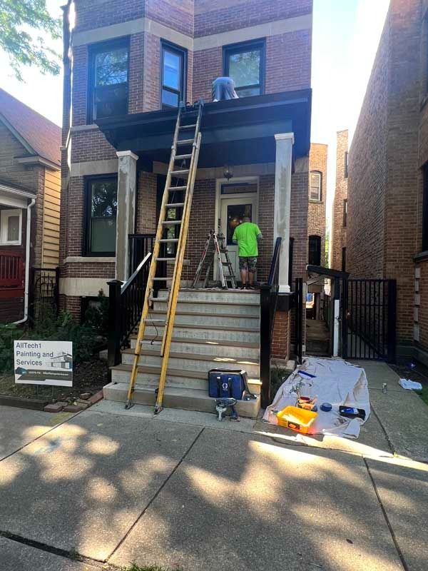Two workers painting the front of a brick building. Ladder propped against the roof, tools on the porch.