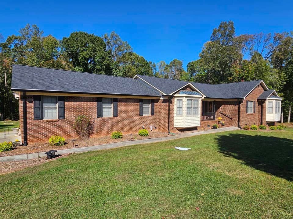 A cream-colored gutter system and downspout installed on the corner of a brick home with an asphalt shingle roof.