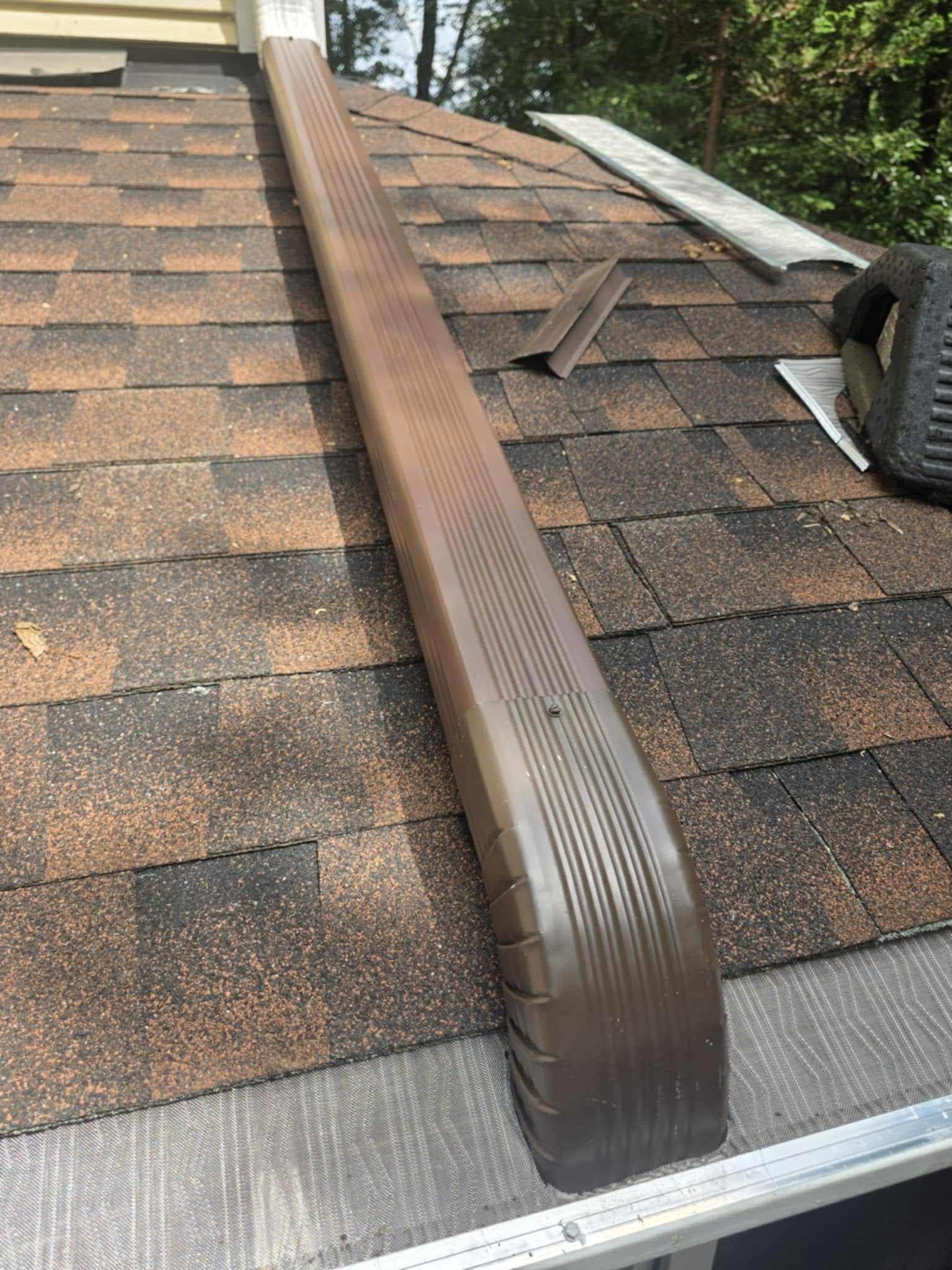 A brown gutter downspout extension sits on a residential shingled roof, leading into a metal gutter below.