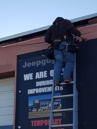 A person wearing a tool belt stands on a ladder, working on a large sign that says 