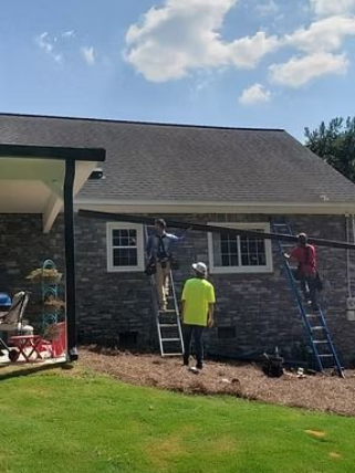 Three workers use ladders to install a long black gutter beam onto the side of a stone-faced house.