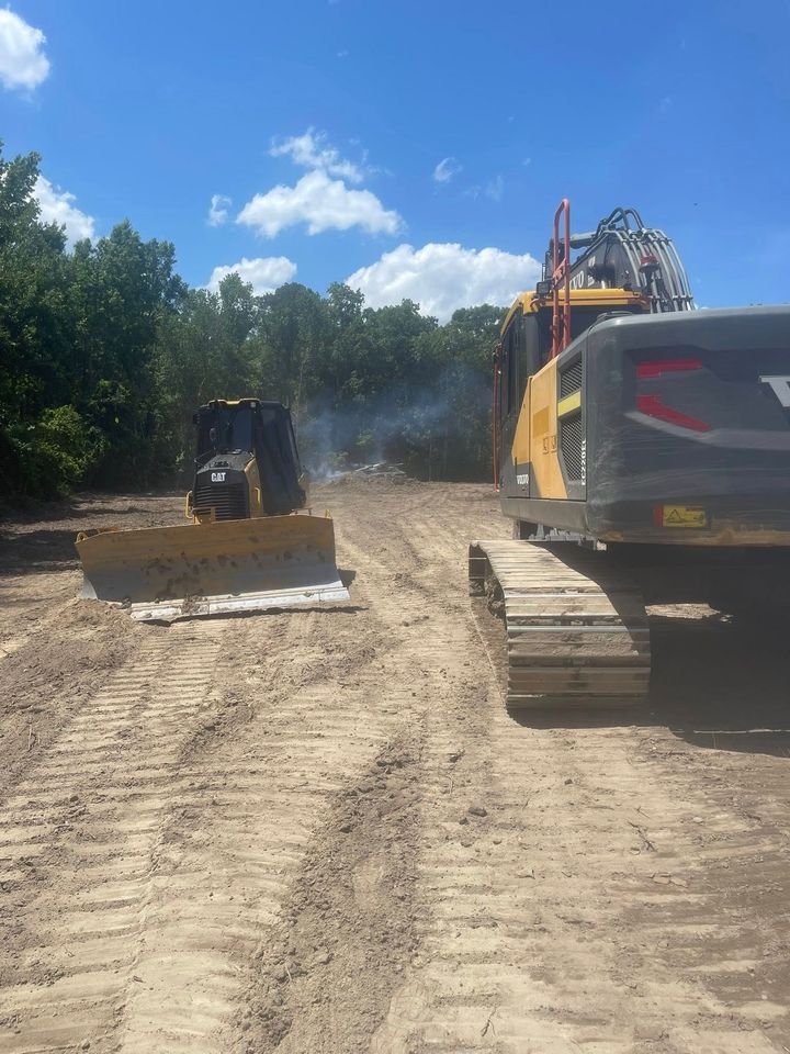 Construction equipment on a dirt road, with a bulldozer and excavator clearing a wooded area under a blue sky