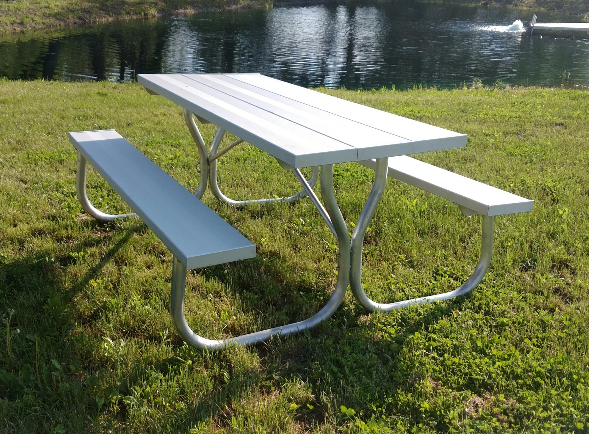 Aluminum picnic table with attached benches on green grass near a pond.