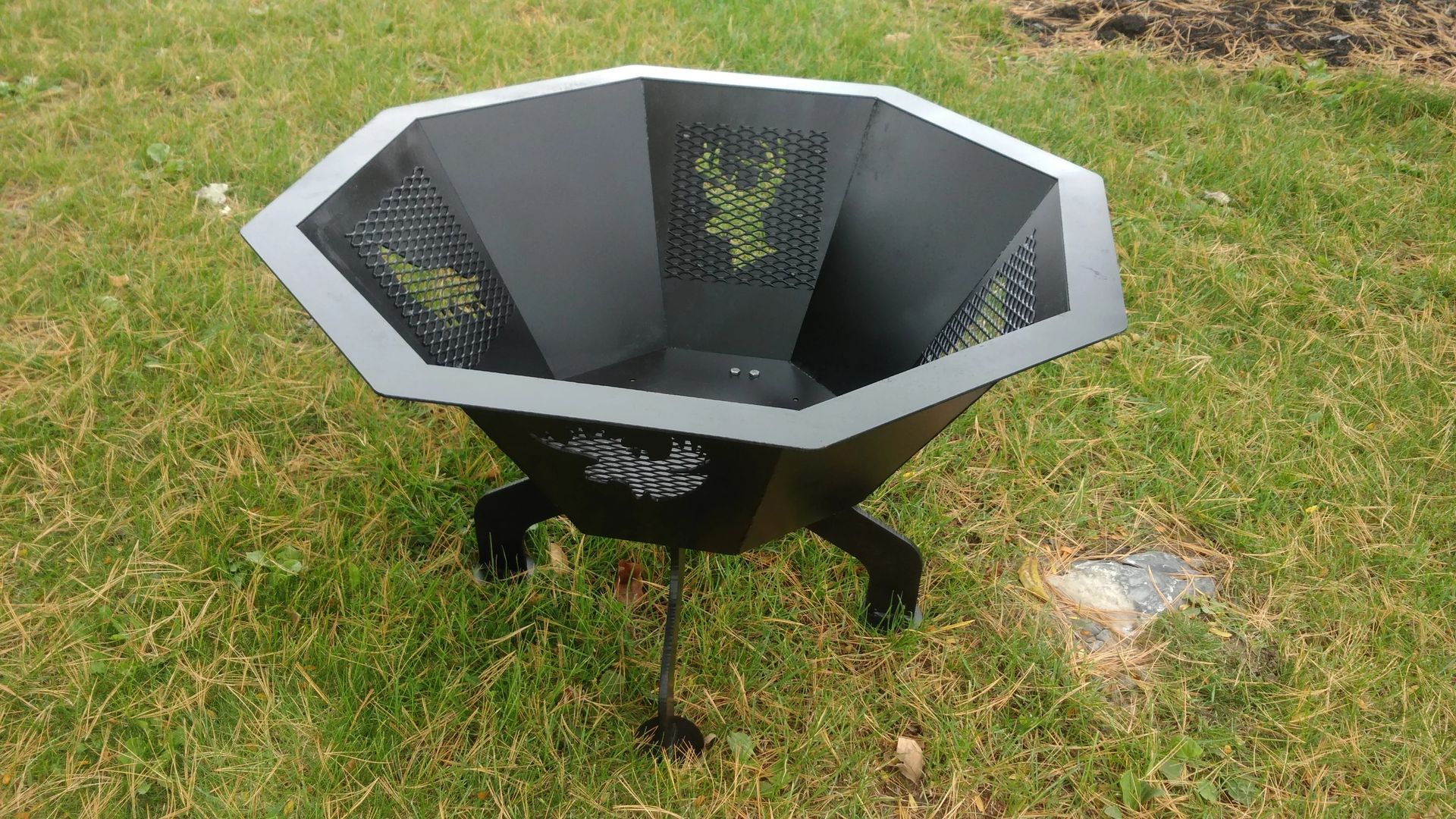 Black metal fire pit with angular design, on green grass.