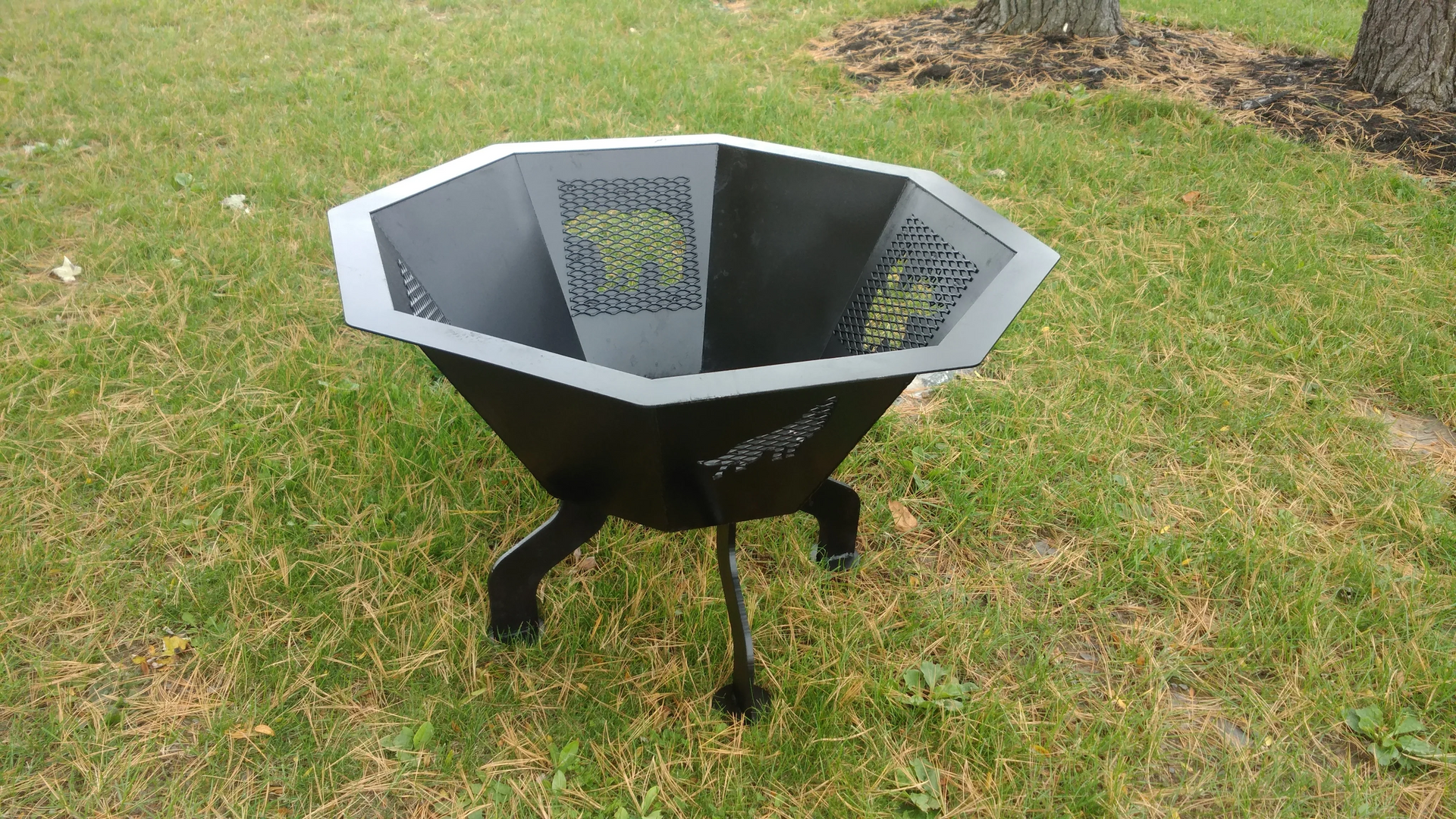 Black, octagonal fire pit on three legs, set on grass lawn.