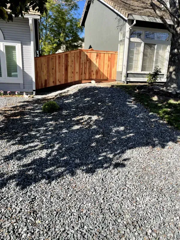 A wooden fence is surrounded by gravel in front of a house.