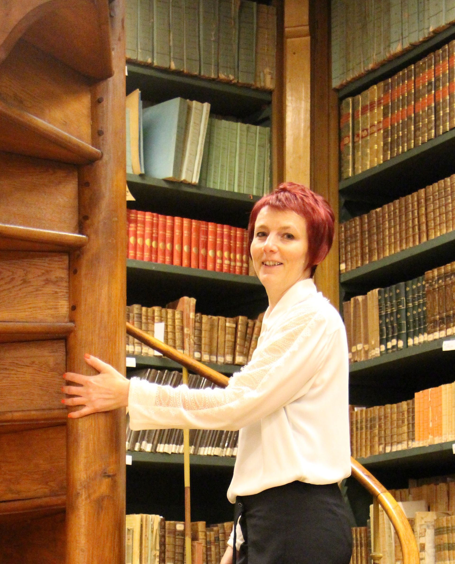 Une femme en chemise blanche se tient à côté d'un escalier dans une bibliothèque