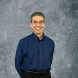 Smiling man in a dark blue shirt standing against a gray textured backdrop