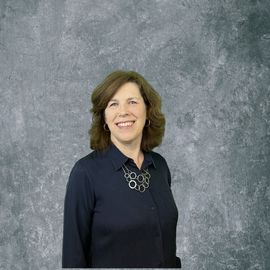 Smiling woman in a dark blouse and necklace against a gray textured backdrop