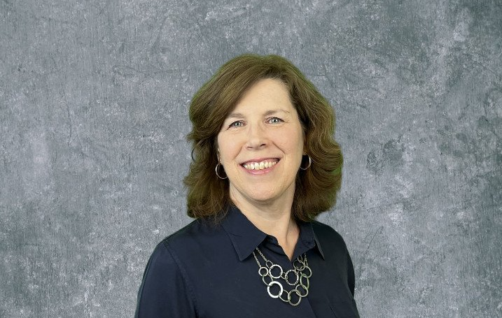 Portrait of a smiling woman in a dark blouse and necklace against a gray textured backdrop
