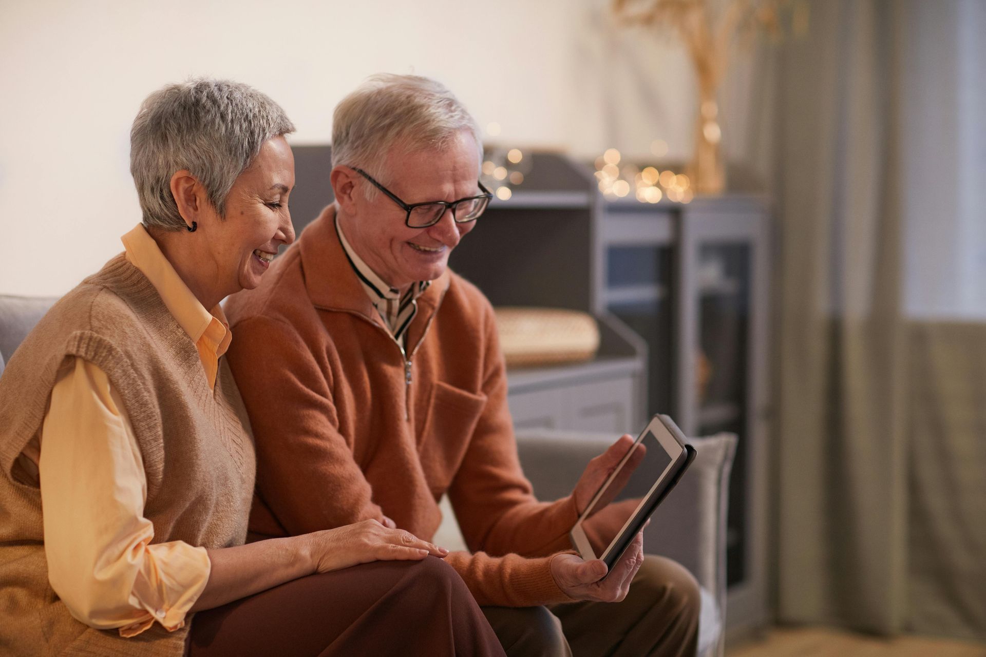 Two older adults sitting on a couch, smiling and looking at a tablet in a cozy living room.